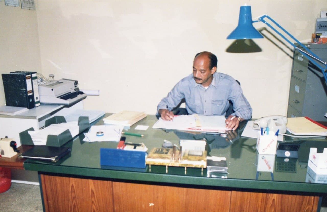 A man writing at a desk under an anglepoise lamp, telephone and ledgers beside him