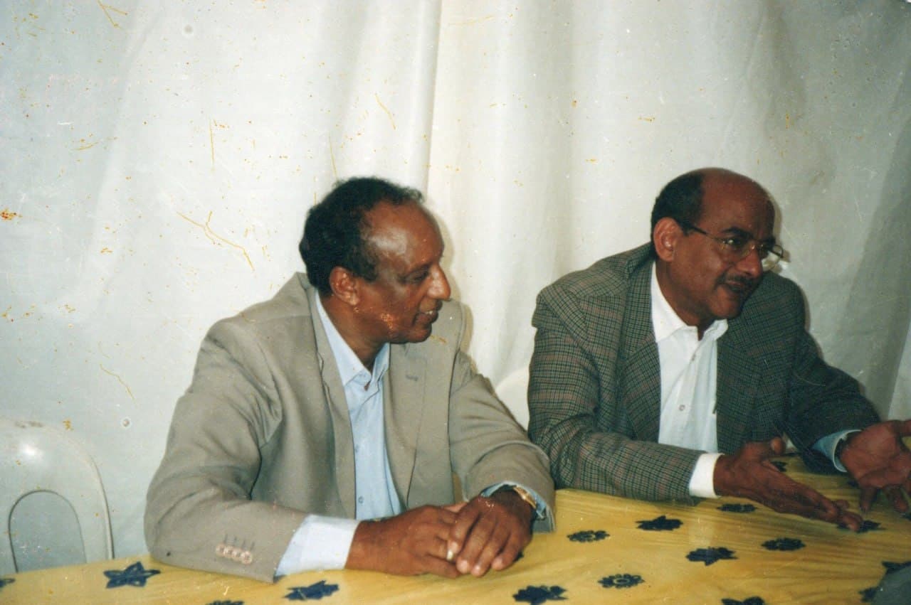 Two men in suits talking across a table, one of them mid-sentence with a hand raised